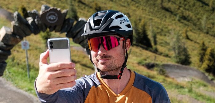 Ein Mann mit Fahrradhelm und Sonnenbrille macht ein Selfie mit seinem Handy.