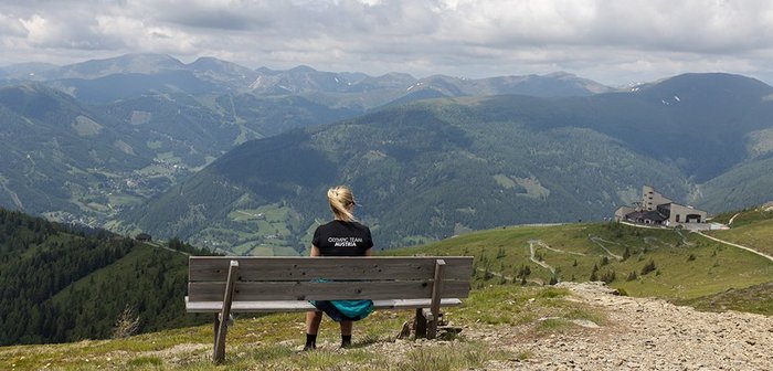 Eine Frau sitzt auf einer Holzbank und beobachtet eine traumhafte Berglandschaft.