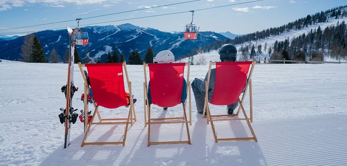 Zwei Erwachsene und ein Kind sitzen auf roten Sonnenstühlen im Schnee.