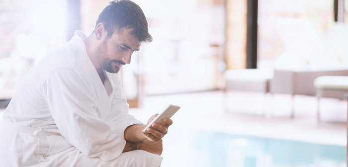 Ein Mann sitzt im Bademantel neben einem Pool und schaut auf sein Handy. 