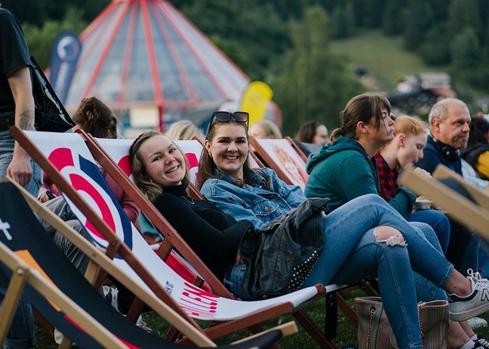 Zwei lachende Frauen sitzen zwischen anderen Personen auf Liegestühlen.