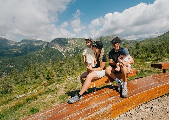 Familie sitzend an der Panoramabank mit Sicht auf die Nockberge, an der Brunnachhöhe