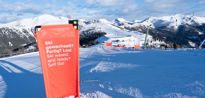 Rotes Schild mit der Aufschrift „Ski gewachst? Fertig? Los!“ auf einer Skipiste inmitten verschneiter Berge.