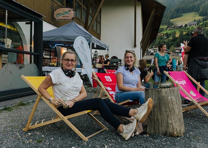 Zwei Frauen mit Kopfhörern um den Hals sitzen auf Liegestühlen.